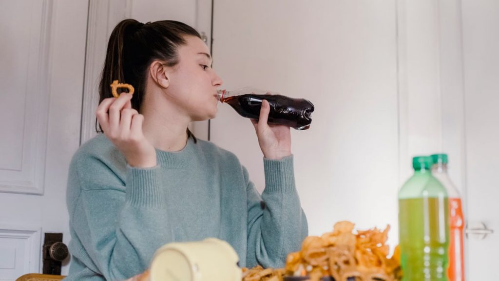 Young female drinking sweet tasty lemonade from plastic bottle at table with different snacks